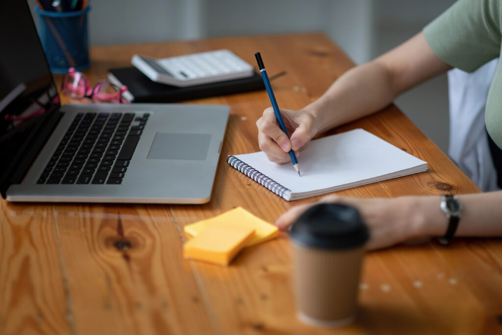 Woman writing a list on a notepad at a desk with a laptop on it.